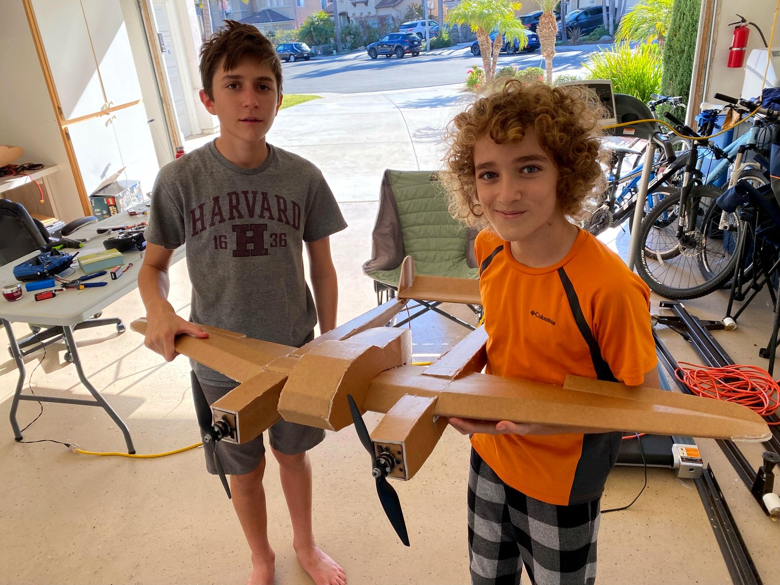 Two boys in a garage holding a hand-built twin-boom, twin-propeller model plane