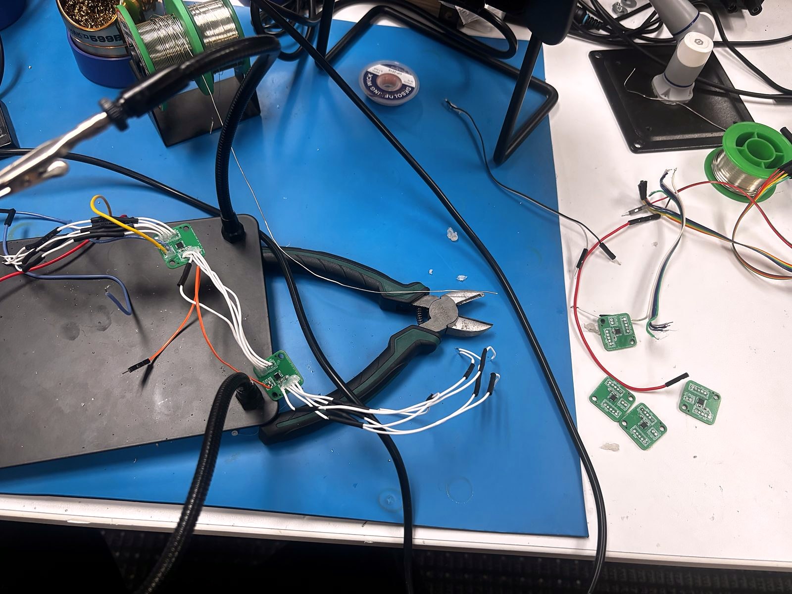 A batch of small square sensor boards on a bench mat, being wired up with ribbon leads next to a soldering station